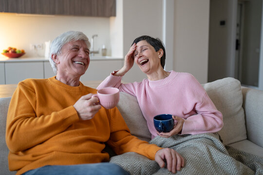 Joyful elderly husband and wife sit on sofa holding colorful cups, laugh during friendly talk, affectionate senior spouses enjoy tea, companionship and cheerful retirement leisure together.
