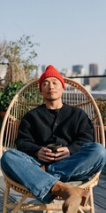 Obraz premium Man meditating on rooftop rattan chair holding a cup wearing a red beanie with skyline view for mindfulness poster and calm background