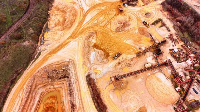 Heavy machinery works a sand quarry site. Trucks and loaders maneuver around swirling tire tracks, conveyor towers and stained earth mounds.