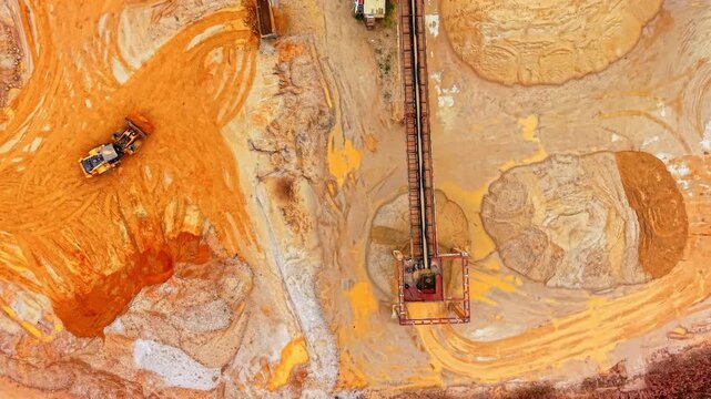 Close aerial view of conveyor tower in sand pit. Loader shifts orange stained sand beside a steel gantry and weathered rust colored terrain.
