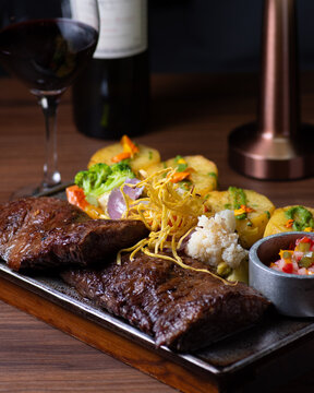 Carne asada a la parrilla servida en fuente de hierro con papas salteadas verduras y copa de vino tinto
