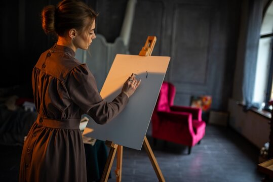 Elegant young woman painting at easel in dim studio with dramatic shadows and focused expression