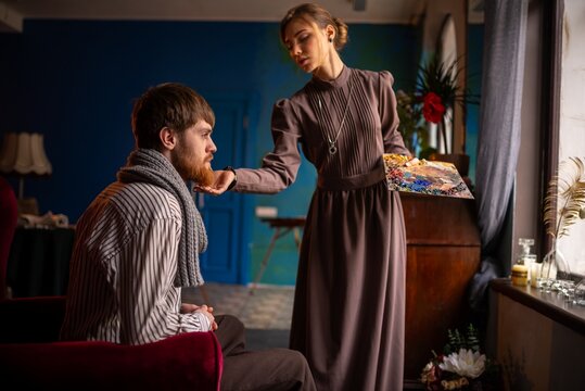 Man seated on upholstered chair facing woman standing beside upright piano in intimate room