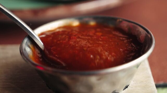 Metal bowl of red sauce being stirred with spoon on wooden table, spicy condiment or salsa, close-up food video footage.