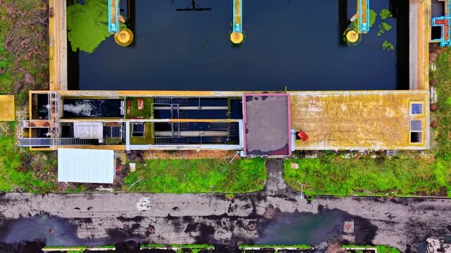 Overhead view of bubbling sedimentation tanks at a treatment plant. Dark water channels, metal grating, pipework and a rooftop storage bin line the facility.