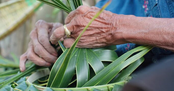 A woman, who is senior, weaves traditional hats from coconut palm leaves by hand on a wooden porch in rural Thailand.