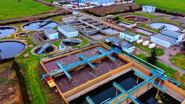 Wastewater treatment facility with clarifiers and support buildings. Circular basins, aeration tanks, access roads and green farmland surround the complex.