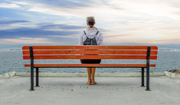 A woman sits alone on a wooden bench facing the sea under a cloudy sky.
