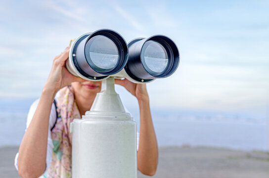 A woman looks through a large stationary binocular telescope outdoors under a bright sky.