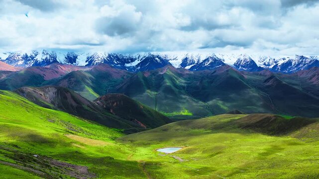 Sunlit alpine grassland rolls toward a small pond beneath a distant snowcapped range, with dramatic clouds casting shadows across the valley.