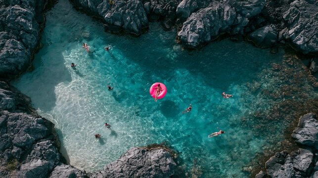 Aerial view of people swimming in a natural rocky sea pool with crystal clear turquoise water and a person floating on a bright pink ring