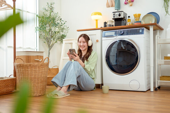 Asian Woman Using Smartphone and Listening to Music While Doing Laundry