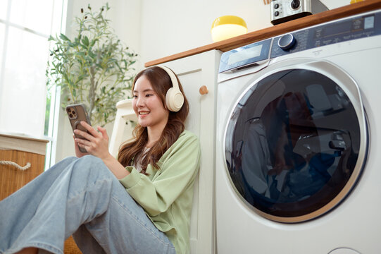 Asian Woman Using Smartphone and Listening to Music While Doing Laundry
