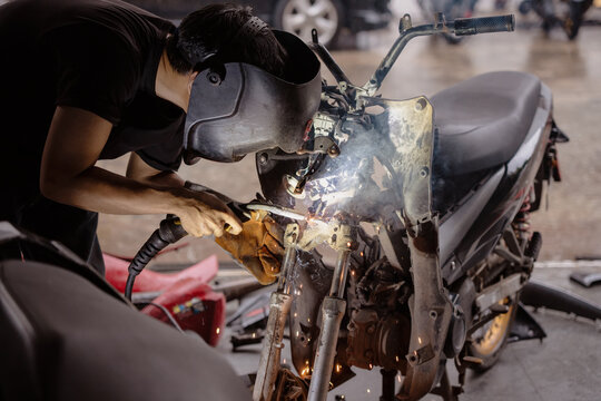 Welder mechanic welding motorcycle steel frame with sparks in custom repair workshop.