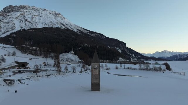 Aerial view of the historic submerged bell tower of the old church rising from the frozen Reschensee lake surrounded by snow-covered mountains in Graun im Vinschgau, Trentino-Alto Adige, Italy.