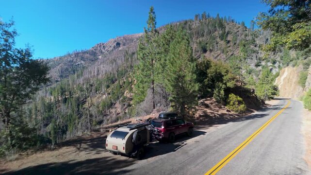 SUV pulls a teardrop camper trailer along a winding road in the Sierra mountains, surrounded by towering trees and scenic views.