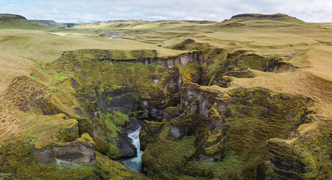 Fjadrargljufur canyon in South Iceland seen from above, with the Fjadra River winding between moss covered cliffs and a grassy plateau under a cloudy summer sky.