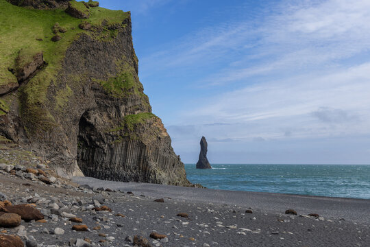 Columnar basalt cliffs rise above the black pebble shore as a Reynisdrangar sea stack stands offshore at Reynisfjara Beach on the Atlantic coast of South Iceland.