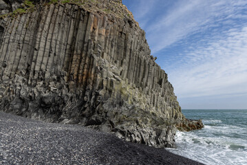 Towering basalt columns rise above black pebbles and Atlantic surf at Reynisfjara Beach in Iceland, revealing rugged volcanic geometry beneath a blue, clouded sky. © Artem