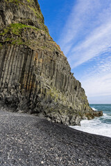 Towering basalt columns rise above the black pebble shore and Atlantic surf at Reynisfjara Beach in Iceland beneath a vivid blue sky with sweeping white clouds. © Artem