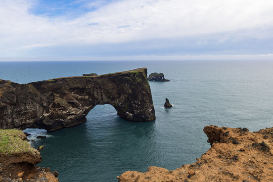 Massive Dyrholaey Arch rises above the ocean beside offshore sea stacks, viewed across rugged foreground cliffs beneath layered clouds on the southern coast of Iceland.