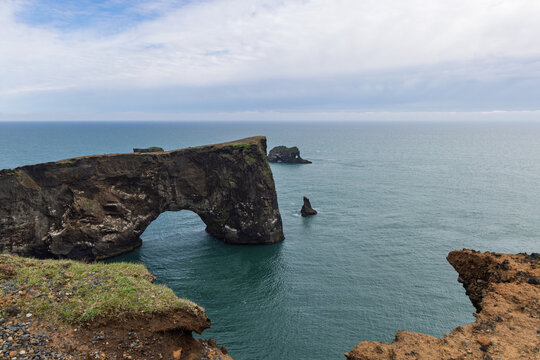 Wide coastal view of Dyrholaey Arch, a massive natural opening in rugged volcanic sea cliffs, with offshore rock formations and the Atlantic Ocean in South Iceland.