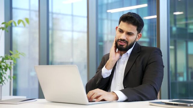 Unwell businessman in formal suit suffering from toothache while sitting at workplace at desk in business office. Male worker holds his hand on his cheek, massaging sore spot, needs to see a dentist.