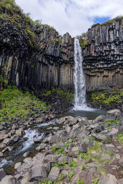 Svartifoss waterfall drops between dark columnar basalt cliffs into a rocky pool in Skaftafell, Iceland, with a flowing stream, scattered boulders and green plants in the foreground.