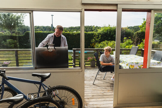 Father barbecuing on patio deck seen through sunroom window