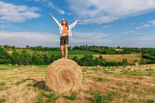 Young woman triumphantly standing atop a round hay bale, arms wide open, peace sign, rolling rural farmland stretching behind her. Adventure concept.
