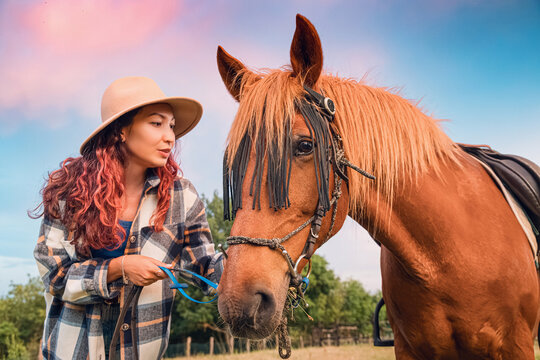 Young woman, gently holding her horse on a ranch with a colorful sky