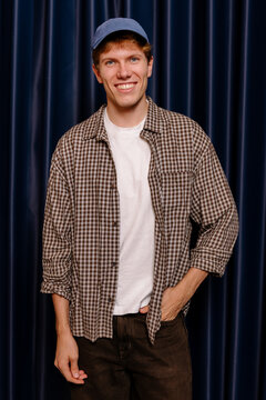 Young man in a cap smiling and posing against a blue curtain.