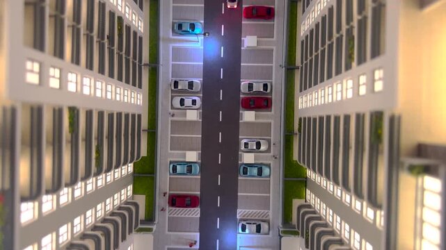 Top down view of an architectural scale model featuring modern residential buildings, parking lots with tiny cars, and realistic illuminated windows in evening. 4K video.