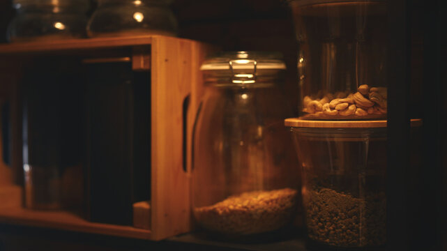 Glass storage jars with cashews, dried chamomile flowers, wooden box and containers on an open kitchen shelf. Cozy home pantry organization in a warm interior.