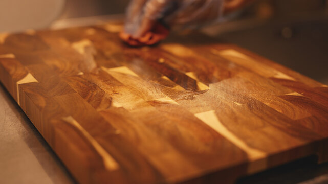 Close-up of a gloved hand rubbing hard wax oil onto an acacia wooden cutting board with a cloth for wood protection and care. This process restores the natural grain texture of the wood surface.