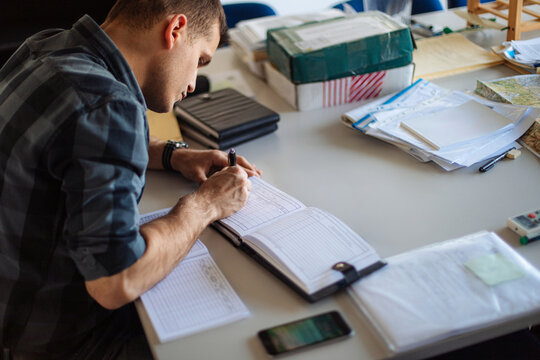 Man Writing in Logbook at Desk With Maps