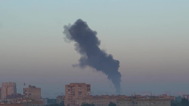 Black smoke column rising over a city. Apartment buildings under a hazy sky. Fire smoke plume above urban skyline.