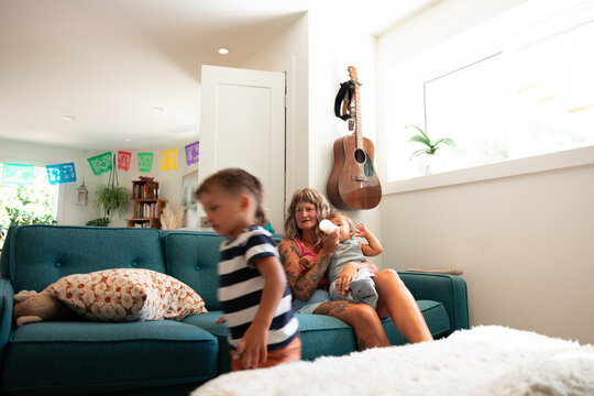 Candid mother feeding baby on couch while toddler moves around