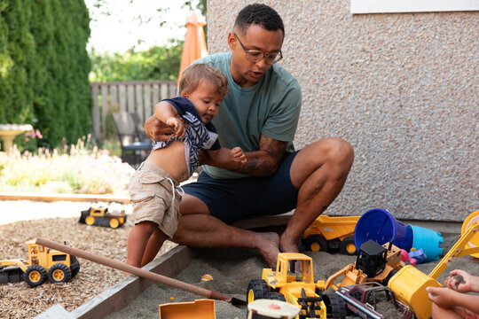 Father helping toddler play with toy trucks in backyard sandbox