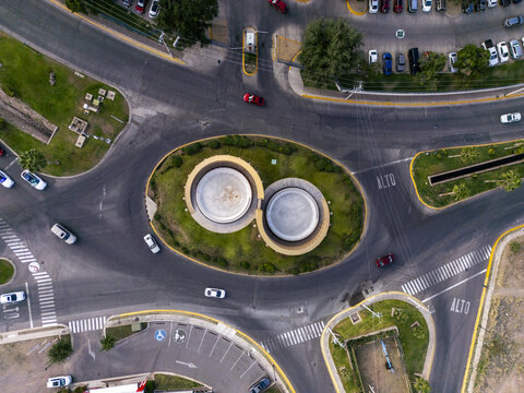Aerial view of a circular roundabout featuring a unique double-circular structure surrounded by green landscaping, roads, and parked cars in Chihuahua, Chihuahua, Mexico.