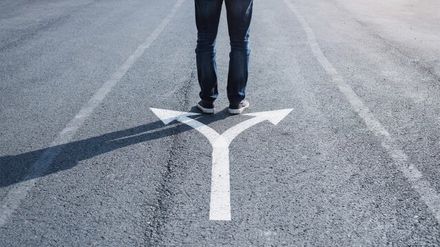 Person standing at a road fork with split white arrows painted on asphalt indicating two different directions for future decisions