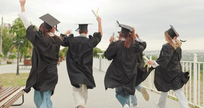 Graduates celebrate graduation while running and jumping outdoors. Footage follows friends in gowns and caps along a park walkway, holding diplomas and raising arms. Pure joy marks a milestone.