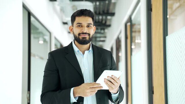 Portrait of smiling businessman in formal suit using a digital tablet and looking at camera in modern business center hallway. Male employee reading message, browsing, chatting online with a client. 