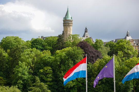 National flags of Luxembourg and the European Union fluttering in the foreground with the clock tower of the historic Spuerkeess building rising above the lush green P&eacute;trusse Valley.