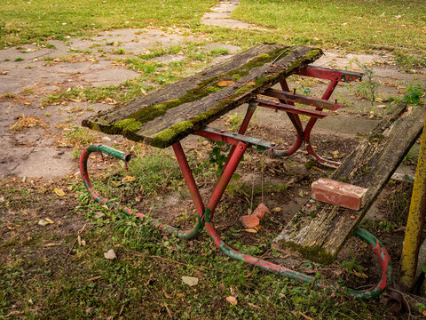 Old broken and decayed wooden picnic table with moss and rust in an abandoned park area overgrown with weeds