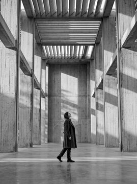 Young woman walking and gazing up inside a monumental concrete colonnade with dramatic sunlight and shadows