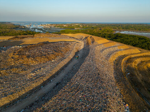 Aerial view of TPA Suwung landfill site with large waste piles, a dirt road, and surrounding mangrove forests near the coast in Kecamatan Denpasar Selatan, Bali, Indonesia.