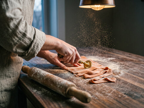 Person cutting homemade fresh pasta dough ribbons with a brass cutter wheel on a rustic wooden table under warm lighting