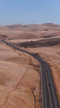 Aerial perspective of vehicles driving along a winding asphalt highway through dry rolling hills near an array of wind turbines in California under daylight.