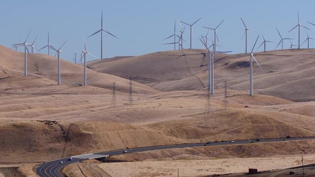 Aerial perspective of modern wind turbines spinning across dry rolling hills near a curving multi lane highway with vehicle traffic in sunny California.